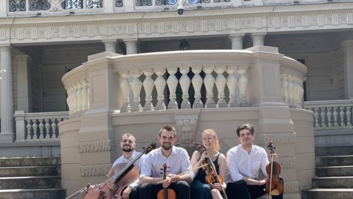 A woman and 3 men – members of the Vivenda String Quartet with string instruments sitting in front of the building