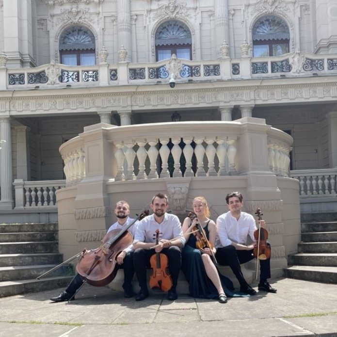 Kobieta i 3 mężczyzn - członkowie zespołu Vivenda String Quartet, siedzą z instrumentami smyczkowymi przed budynkiem