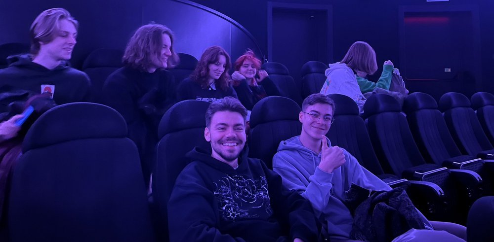 Zdjęcie przedstawia młode osoby siedzące w fotelach kinowych przy ciemnoniebieskim świetle.