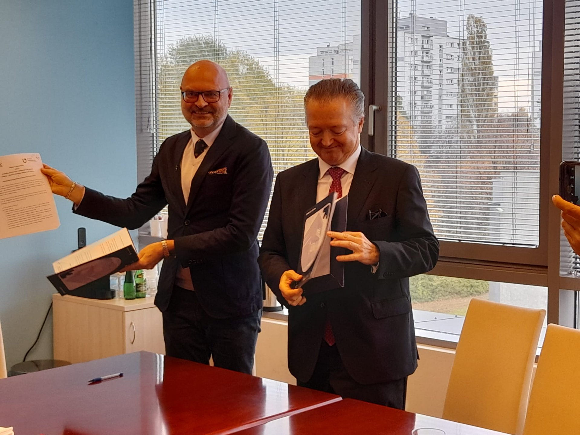 Dwie osoby w formalnym ubiorze siedzą przy biurowym stole, podpisując dokumenty. Jedna osoba ma łysą głowę i nosi okulary, druga ma gęste włosy zaczesane do tyłu. Za nimi widać jasne okno, przez które wpada światło dzienne.