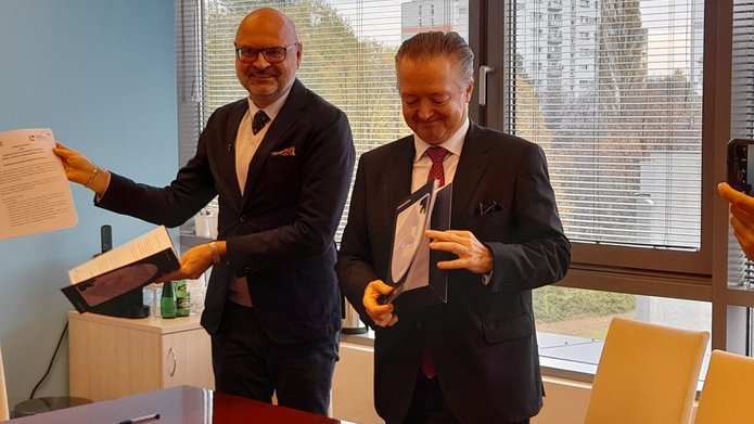 Dwie osoby w formalnym ubiorze siedzą przy biurowym stole, podpisując dokumenty. Jedna osoba ma łysą głowę i nosi okulary, druga ma gęste włosy zaczesane do tyłu. Za nimi widać jasne okno, przez które wpada światło dzienne.