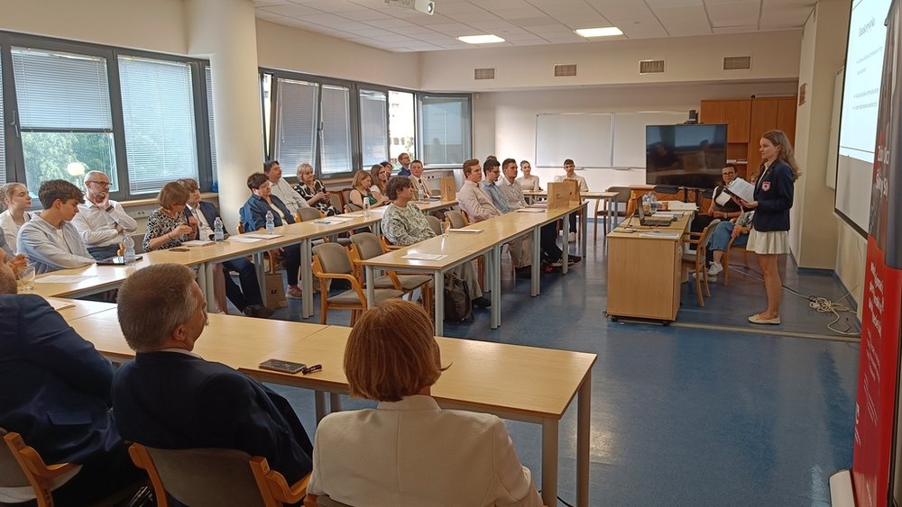 Na zdjęciu znajduje się sala konferencyjna wypełniona uczestnikami spotkania. Ustawienie stołów w kształt litery "U". Prelegentka stoi przed ekranem prezentacyjnym po prawej stronie. To młoda kobieta, ubrana w jasny strój, przedstawia slajdy z prezentacji. Sala jest dobrze oświetlona, z dużymi oknami po lewej stronie wpuszczającymi naturalne światło. Uczestnicy siedzą na krzesłach przy stołach, słuchając prelegentki. Po prawej stronie, na ścianie, widoczny jest baner z logo Zdolny uczeń-świetny student”.