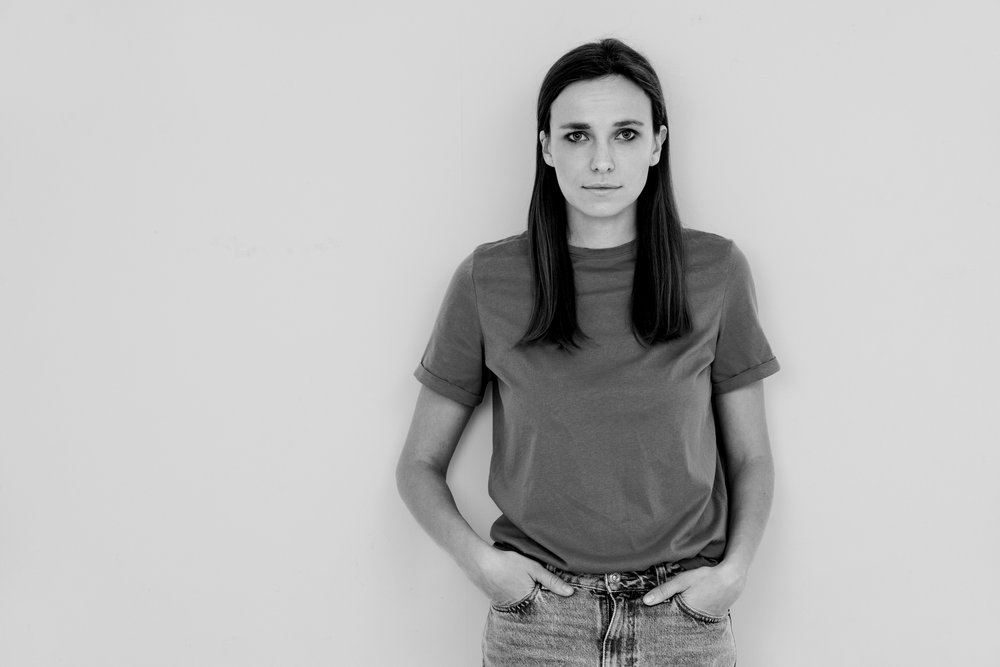 A black and white photo of a young woman with long hair leaning against a white wall