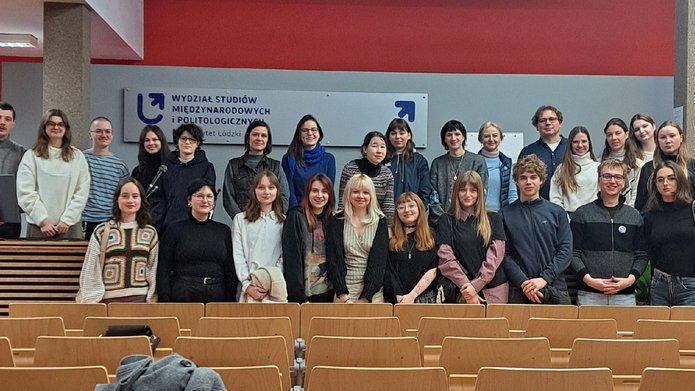 Zdjęcie grupowe wykonane w sali wykładowej Wydziału Studiów Międzynarodowych i Politologicznych Uniwersytetu Łódzkiego. Przed tablicą z nazwą wydziału stoi 26 osób w dwóch rzędach — pierwszy rząd przed ławkami, drugi za nimi. Uczestnicy pozują do wspólnego zdjęcia.