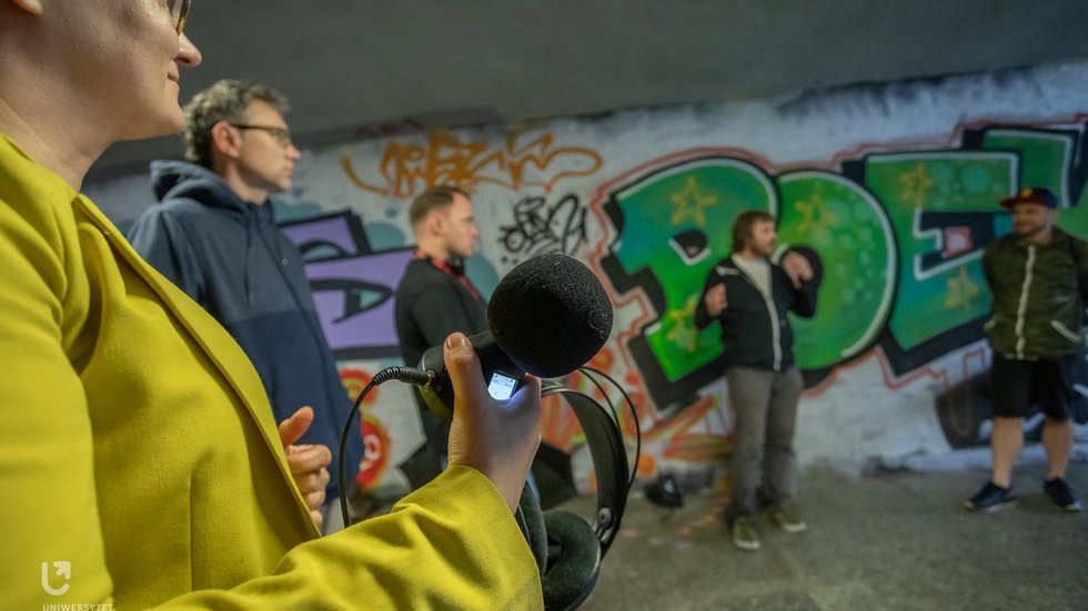 The photo shows a group of people standing in an underpass. They are holding audio recording devices.