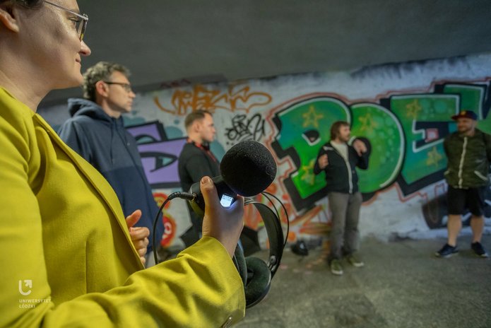 Na zdjęciu widać grupę ludzi. Stoją przejściu podziemnym. Trzymają w rękach urządzenia do nagrywania dźwięków. 