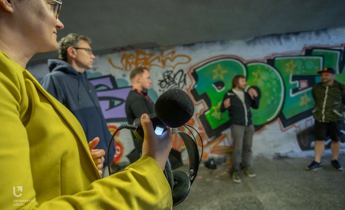 The photo shows a group of people standing in an underpass. They are holding audio recording devices.
