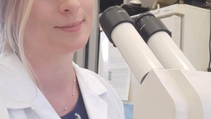 Kolorowe zdjęcie przedstawiające Martę Rudną: doktorantkę Szkoły Doktorskiej Nauk Ścisłych i Przyrodniczych UŁ. Fotografia ukazuje młodą kobietę o blond włosach i niebieskich oczach pozującą z z nowoczesnym mikroskopem ubraną w biały fartuch laboratoryjny
