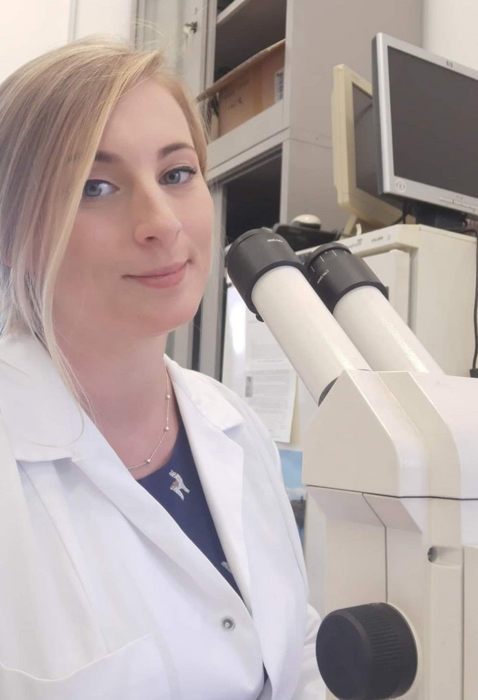 Kolorowe zdjęcie przedstawiające Martę Rudną: doktorantkę Szkoły Doktorskiej Nauk Ścisłych i Przyrodniczych UŁ. Fotografia ukazuje młodą kobietę o blond włosach i niebieskich oczach pozującą z z nowoczesnym mikroskopem ubraną w biały fartuch laboratoryjny