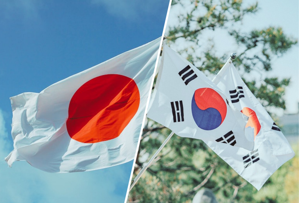 Po lewej powiewająca, biało-czerwona flaga Japonii na tle nieba, po prawo dwie flagi Korei Południowej powiewające na wietrze na tle zielonych liści drzew i błękitnego nieba.