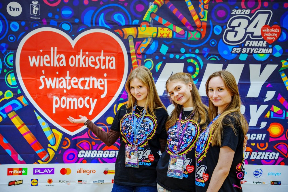 3 młode kobiety w koszulkach z napisem Wielka Orkiestra Świątecznej Pomocy stojące na tle kolorowej grafiki z napisem Wielka Orkiestra świątecznej Pomocy
