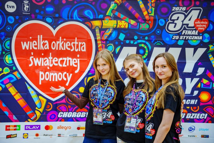 3 młode kobiety w koszulkach z napisem Wielka Orkiestra Świątecznej Pomocy stojące na tle kolorowej grafiki z napisem Wielka Orkiestra świątecznej Pomocy