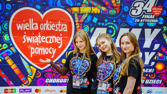 3 młode kobiety w koszulkach z napisem Wielka Orkiestra Świątecznej Pomocy stojące na tle kolorowej grafiki z napisem Wielka Orkiestra świątecznej Pomocy