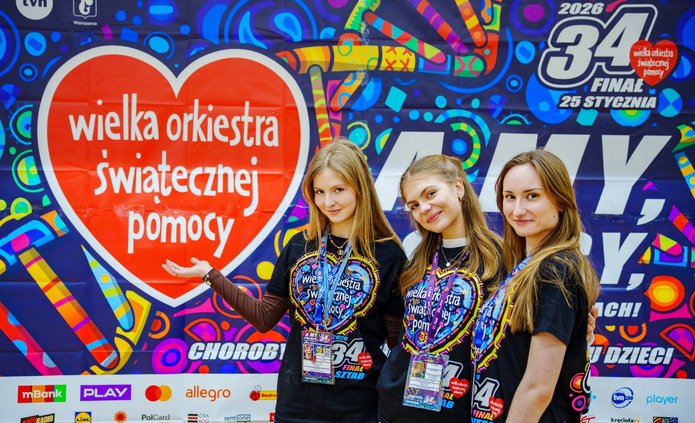 Three young women wearing T-shirts with the words "Great Orchestra of Christmas Charity" written on them standing in front of a colourful graphic with the words "Great Orchestra of Christmas Charity" written on it.