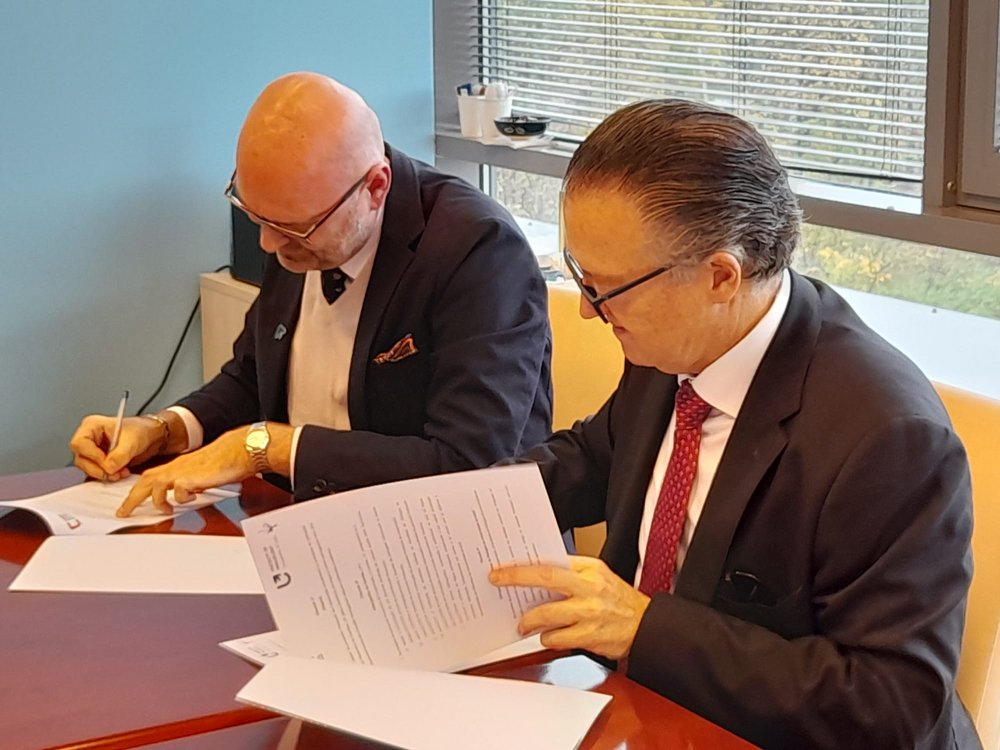 Te same osoby stoją i trzymają dokumenty, wydają się być zadowolone z ich podpisania. Osoba po lewej trzyma dokument w jednej ręce i ma drugą rękę umieszczoną w kieszeni, natomiast osoba po prawej trzyma dokument w jednej ręce i gestykuluje drugą. W tle widoczne są duże okna i jasne ściany pomieszczenia.