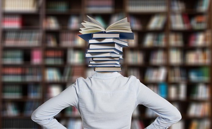 Na zdjęciu widać kobietę od pasa w górę stojącą tyłem plecami. Zamiast głowy ma stos książek
