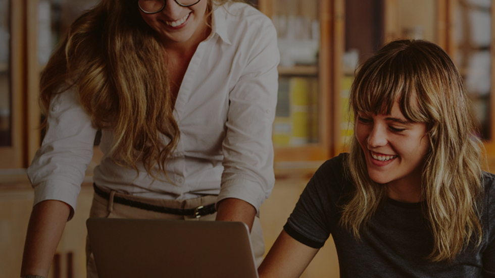 dwie uśmiechnięte kobiety przy biurku spoglądają na laptopa