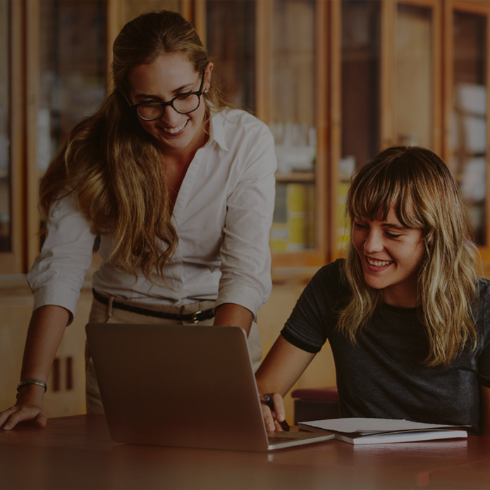 dwie uśmiechnięte kobiety przy biurku spoglądają na laptopa