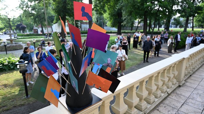Na zdjęciu widać park, w którym jest dużo ludzi. Jest wiosna, drzewa są zielone. Na pierwszym planie widoczna balustrada z betonu na której stoi stojak z kolorowymi chorągiewkami. W tle chodzą wspomniani ludzie.
