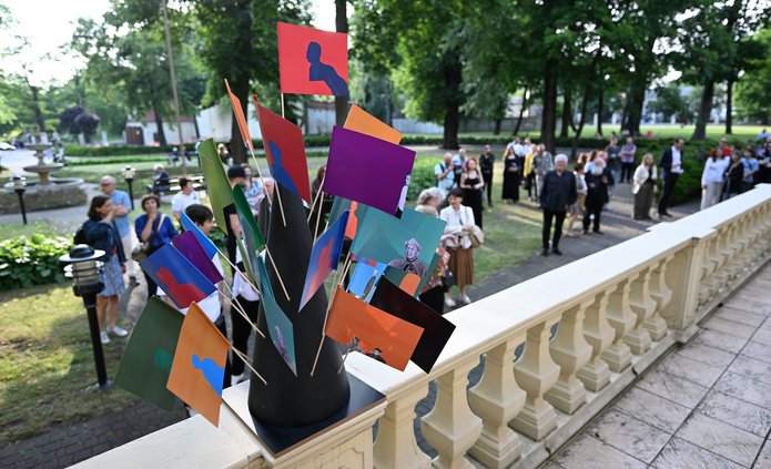 The photograph shows a park filled with people. It is spring, and the trees are green. In the foreground, a concrete balustrade is visible, with a stand displaying colourful bunting placed on top. In the background, the aforementioned people can be seen walking.