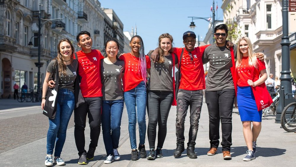 A group of smiling young people with different skin colours wearing University of Lodz T-shirts stand on Piotrkowska Street in Lodz on a sunny day