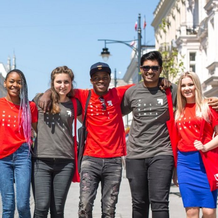 A group of smiling young people with different skin colours wearing University of Lodz T-shirts stand on Piotrkowska Street in Lodz on a sunny day
