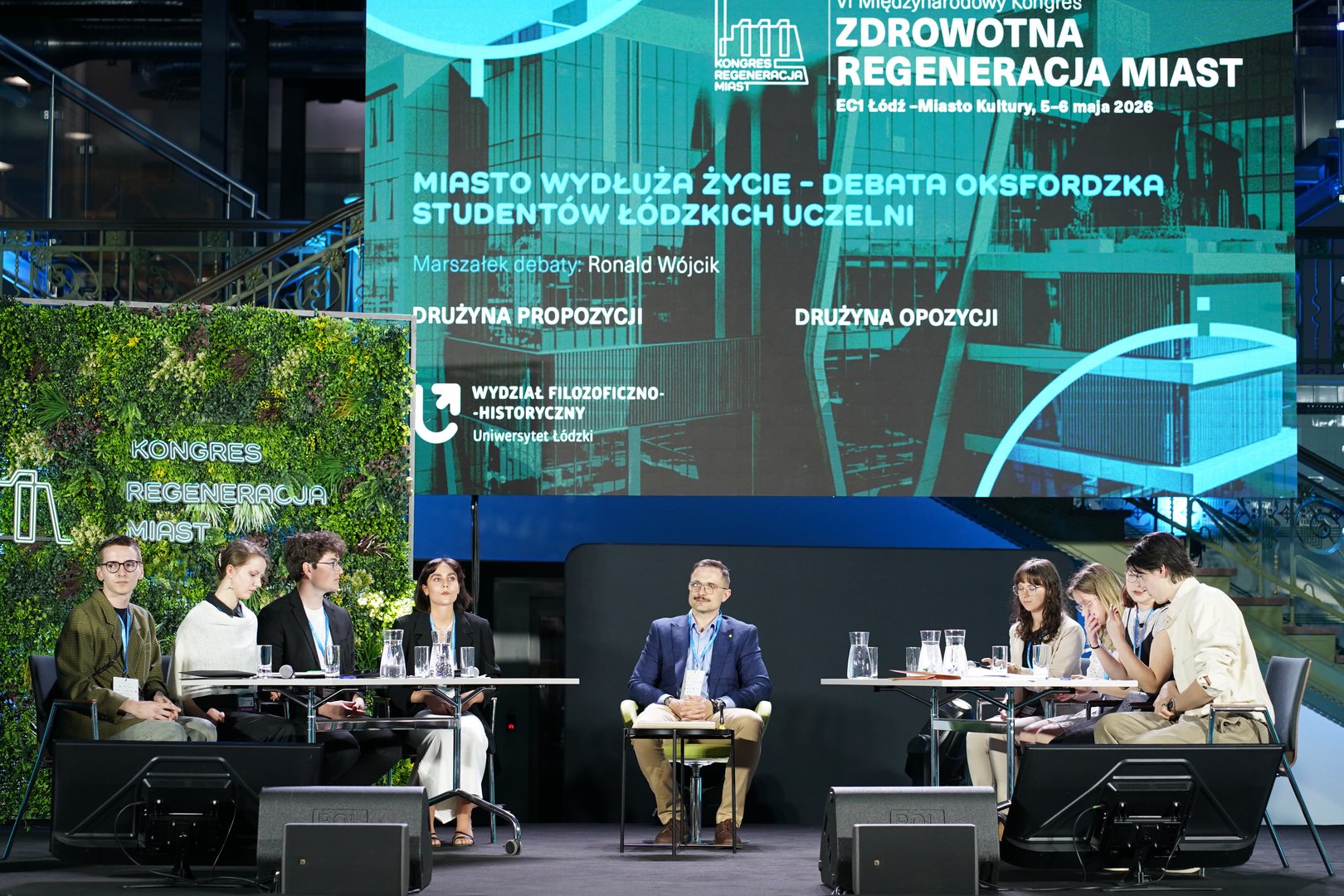 A view of the stage with the debating teams and a large conference screen in the background