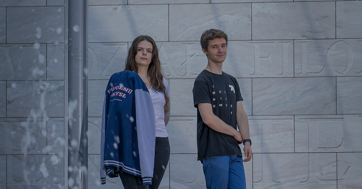 Studentka i student na tle szarej ściany z widocznymi wyrytymi literami. Na pierwszym planie tryskająca woda z fontanny.