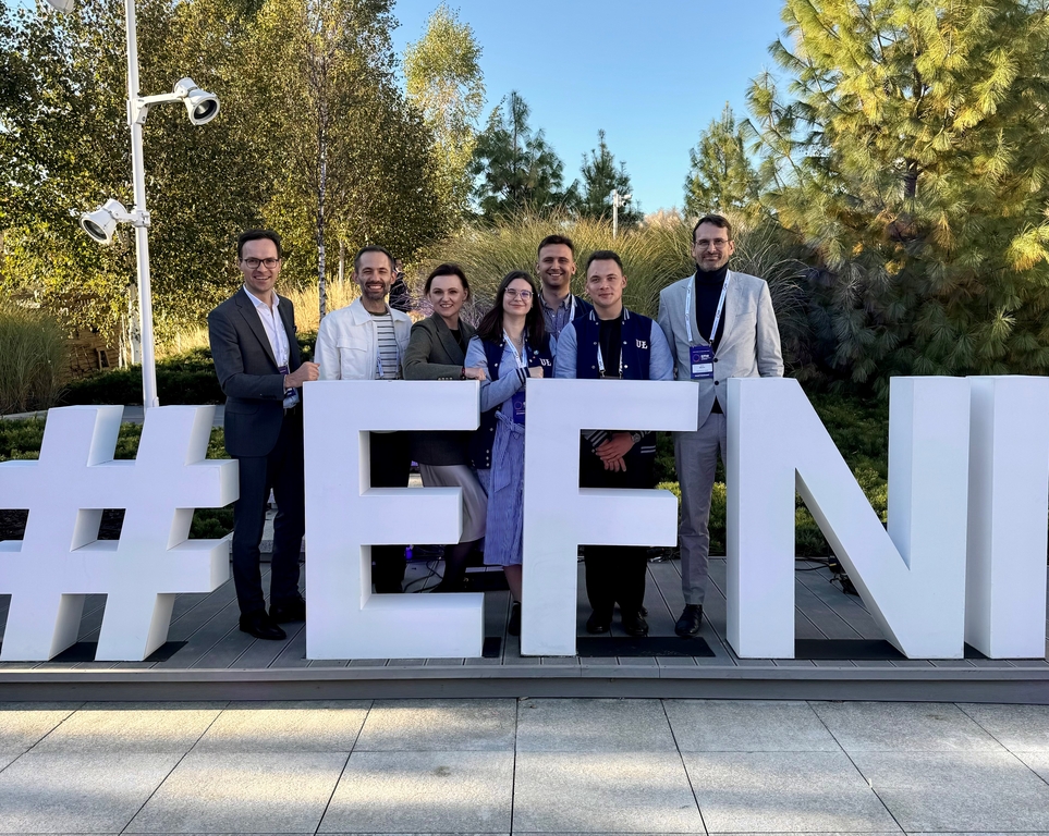 Przedstawiciele Uniwersytetu Łódzkiego, w tym studenci i rektor, stoją przy dużym znaku EFNI, w tle widoczne są drzewa.