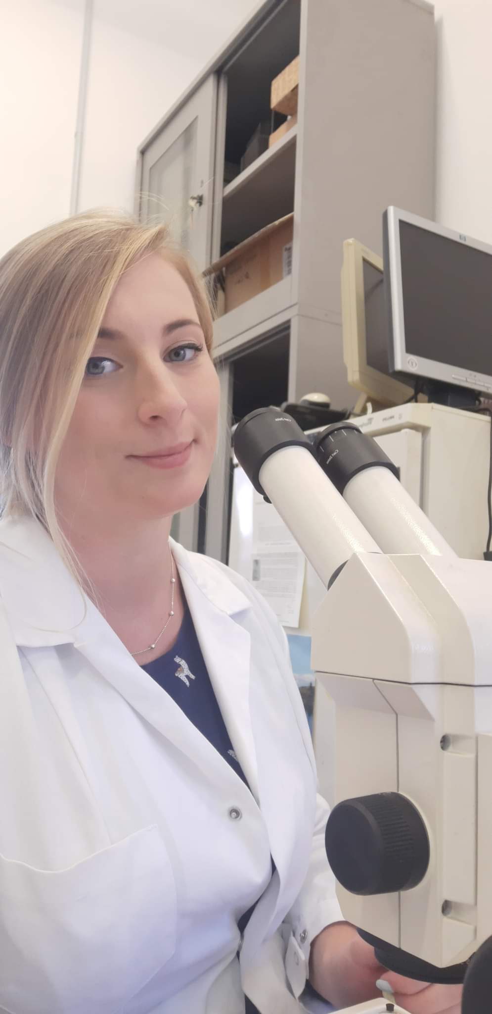 Kolorowe zdjęcie przedstawiające Martę Rudną: doktorantkę Szkoły Doktorskiej Nauk Ścisłych i Przyrodniczych UŁ. Fotografia ukazuje młodą kobietę o blond włosach i niebieskich oczach pozującą z z nowoczesnym mikroskopem ubraną w biały fartuch laboratoryjny