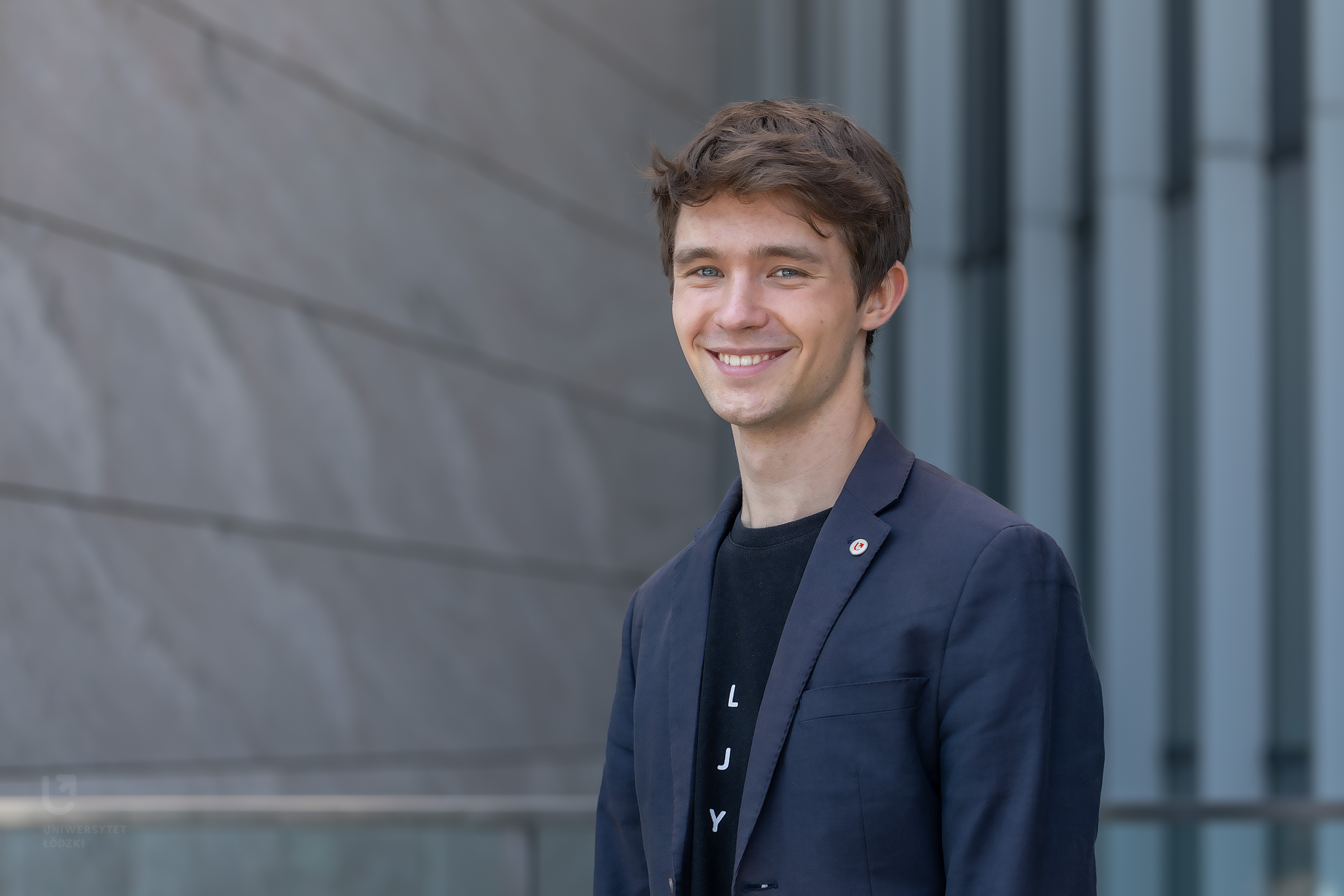 Kolorowe zdjęcje przedstawia Jarosława Brodeckiego: studenta Uniwersytetu Łódzkiego. Student stoi na tle jednego z budynków Uniwersytetu. Jest uśmiechnięty, bardzo przystojny, ubrany w granatową marynarkę.