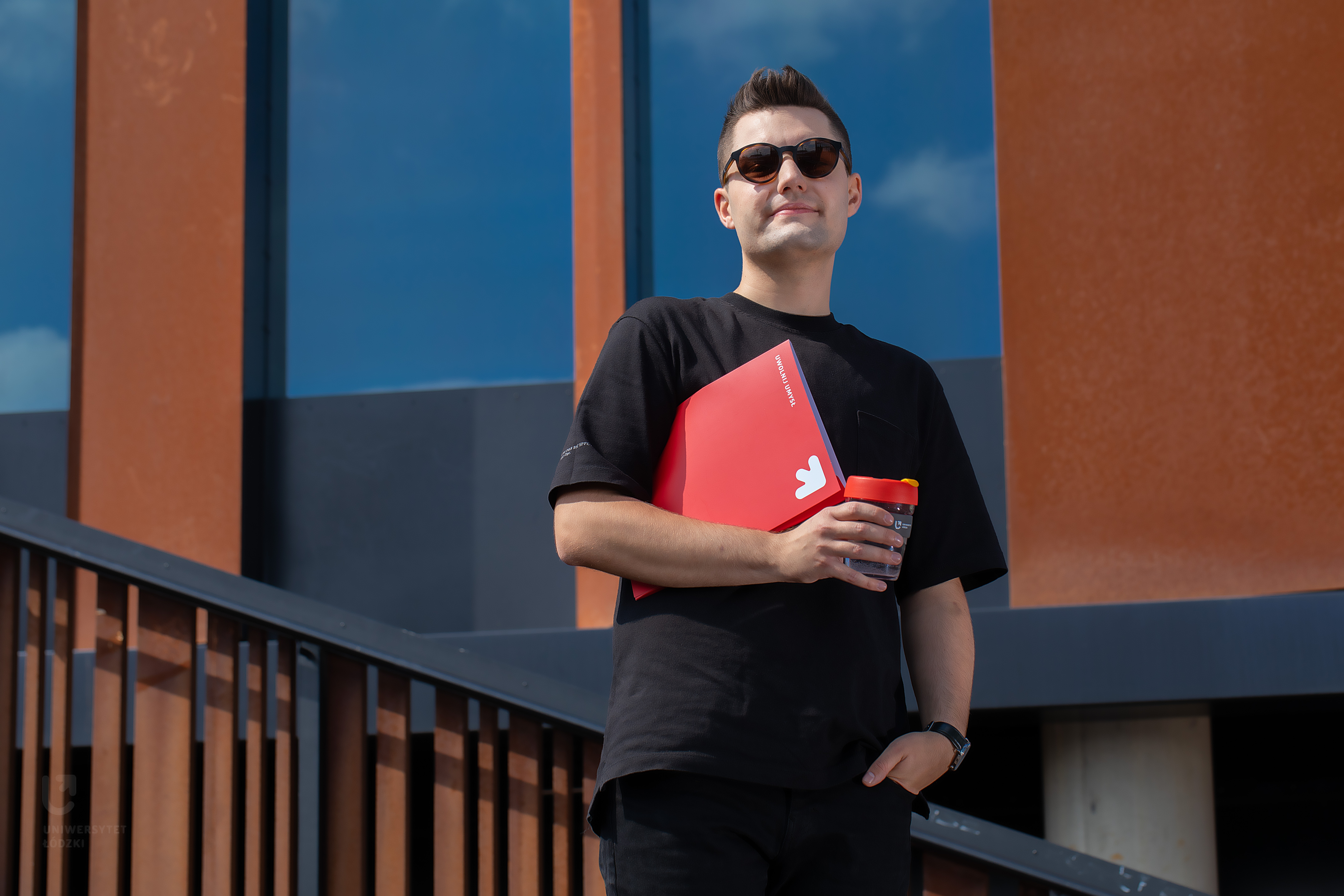 Student w okularach przeciwsłonecznych stojący z czerwoną teczką i kubkiem