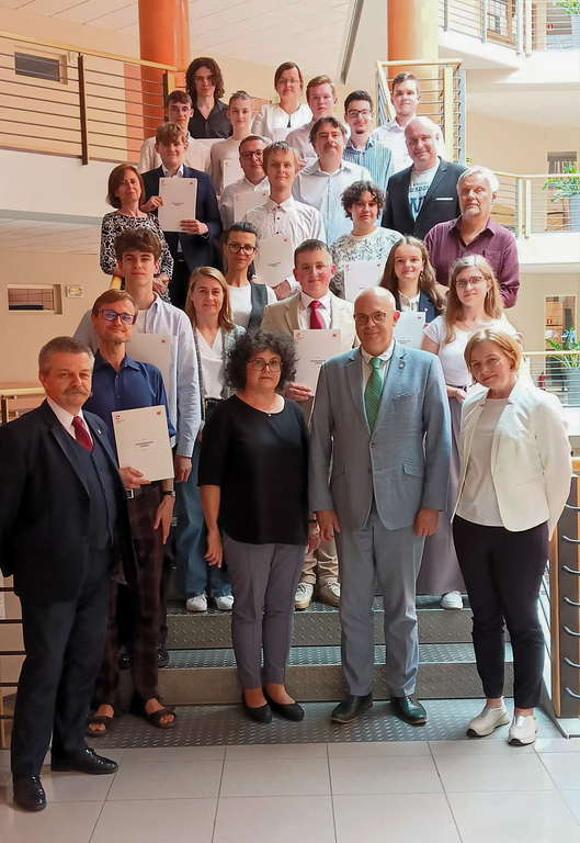 Na zdjęciu znajduje się duża grupa osób, stojących na schodach wewnątrz budynku. Większość z nich trzyma certyfikaty i się uśmiecha. Osoby w grupie są elegancko ubrane. Na pierwszym planie, po prawej stronie, znajduje się mężczyzna w zielonym garniturze oraz kobieta z ciemnymi, kręconymi włosami ubrana na czarno. Wszyscy pozują na tle wnętrza budynku z balustradami i otwartą przestrzenią. 