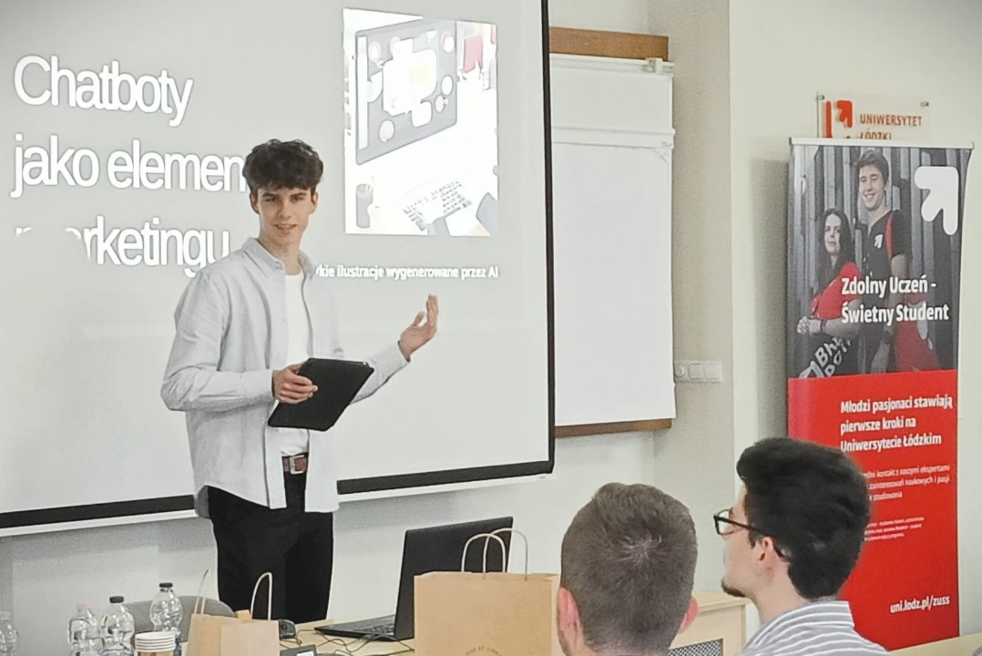 Na zdjęciu znajduje się młody mężczyzna przed ekranem projekcyjnym, na którym wyświetlany jest slajd z napisem "Chatboty jako element marketingu". Prezenter trzyma tablet w jednej ręce i wskazuje na ekran drugą ręką. W tle, obok ekranu, znajduje się baner z napisem „Zdolny uczeń-świetny student”.