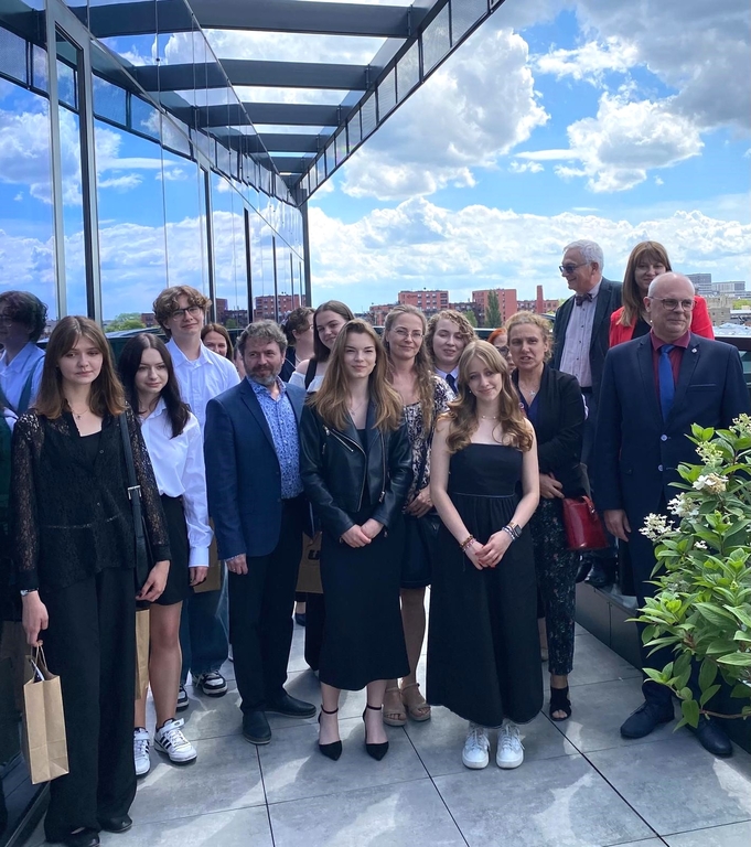 Grupa osób stojących na zewnątrz na tarasie lub balkonie budynku. Wszyscy są elegancko ubrani. Wśród grupy znajduje się zarówno młodzież, jak i dorośli. Niektóre osoby trzymają torby z logo Uniwersytetu Łódzkiego. W tle widać fragmenty miejskiego krajobrazu oraz zieleń.