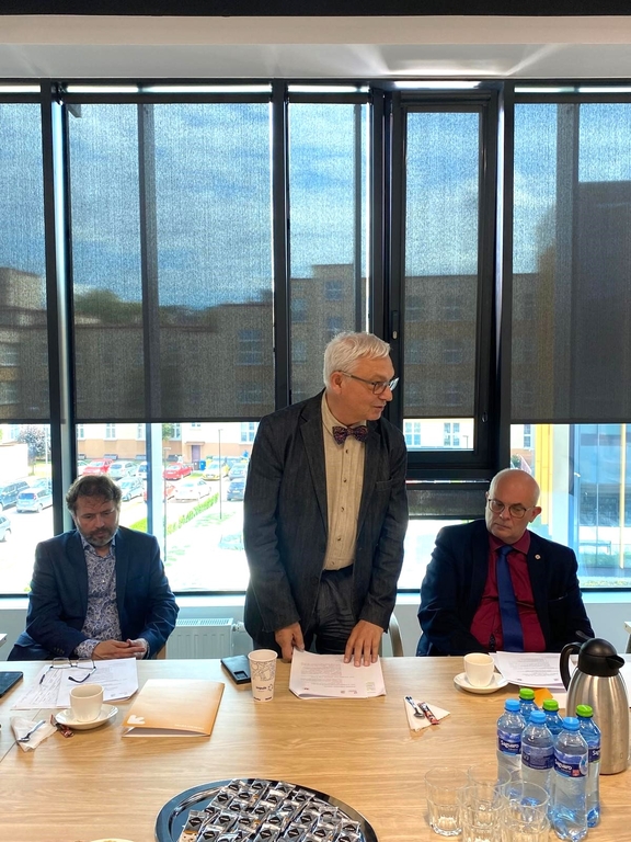 Trzech mężczyzn w sali konferencyjnej. Dwóch z nich siedzi przy stole, a trzeci stoi pośrodku, przemawiając. Mężczyzna stojący, ubrany w szary garnitur i muszkę, trzyma dokumenty i wydaje się być w trakcie przemowy.. Na stole przed nimi znajdują się filiżanki, butelki z wodą, dokumenty i inne przedmioty. W tle widać duże okna