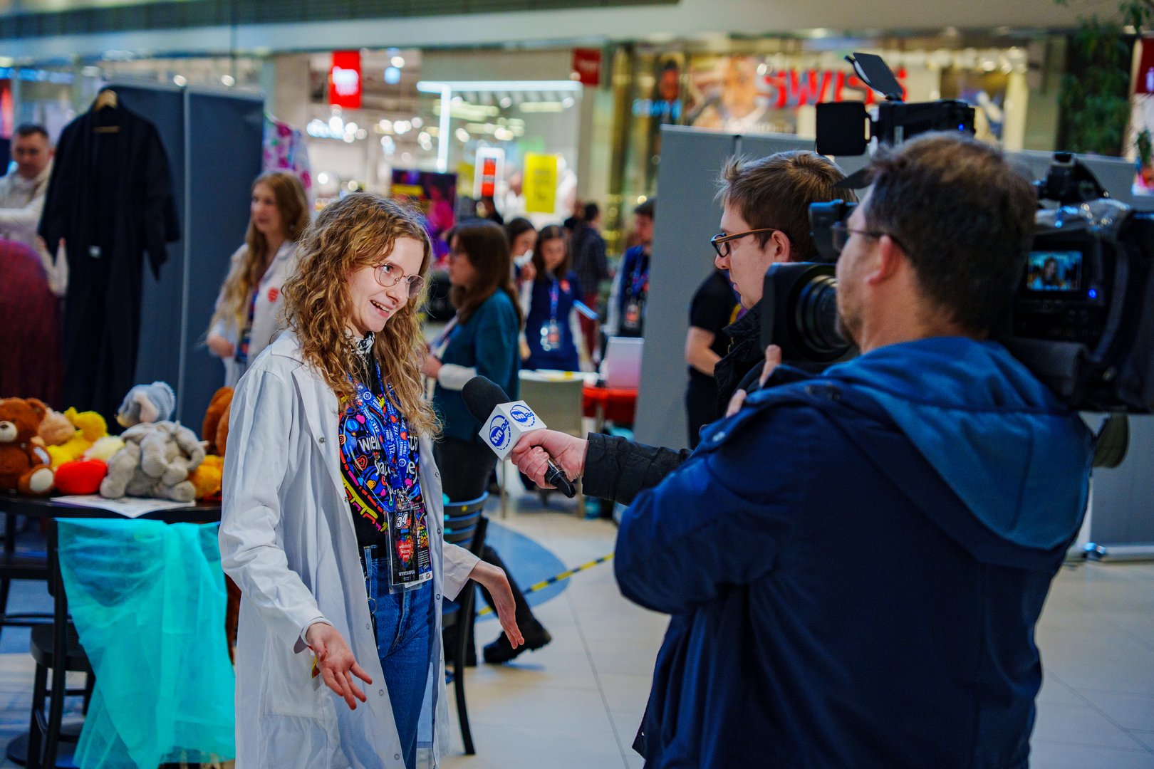 Młode kobieta w koszulce z napisem Wielka Orkiestra świątecznej Pomocy i białym fartuchu stojąca przed 2 dziennikarzami z kamerą i mikrofonem