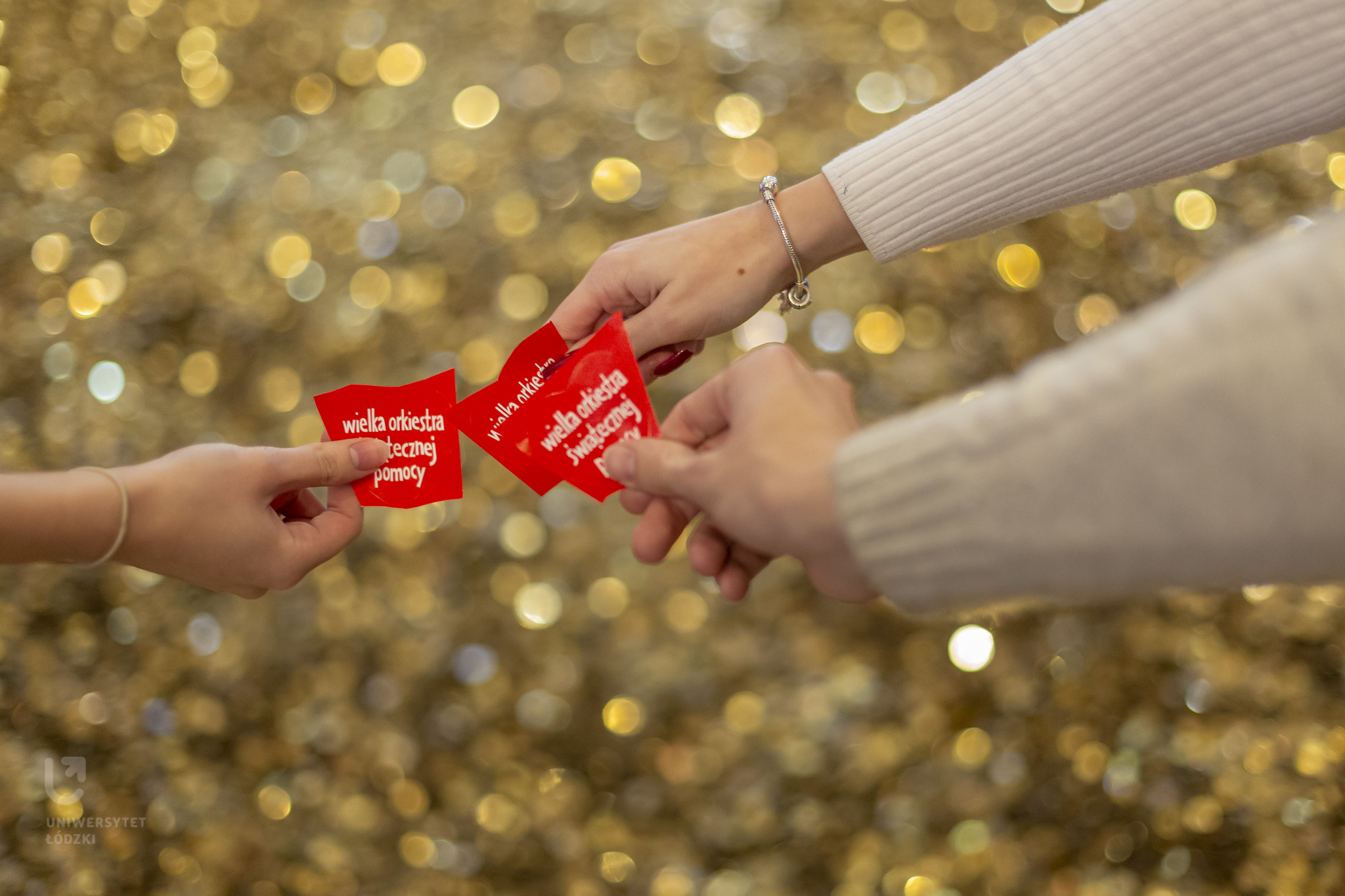 3 ręce trzymające czerwone serduszka z napisem Wielka Orkiestra Świątecznej Pomocy" na tle złotych monet