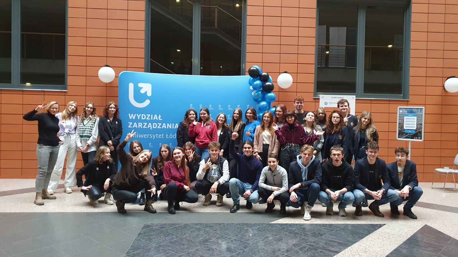 Grupa trzydziestu maturzystów (chłopcy i dziewczęta) stojący przez ścianką Wydziału Zarządzania. Ścianka jest koloru błękitnego z białymi logotypami Wydziału Zarządzania.