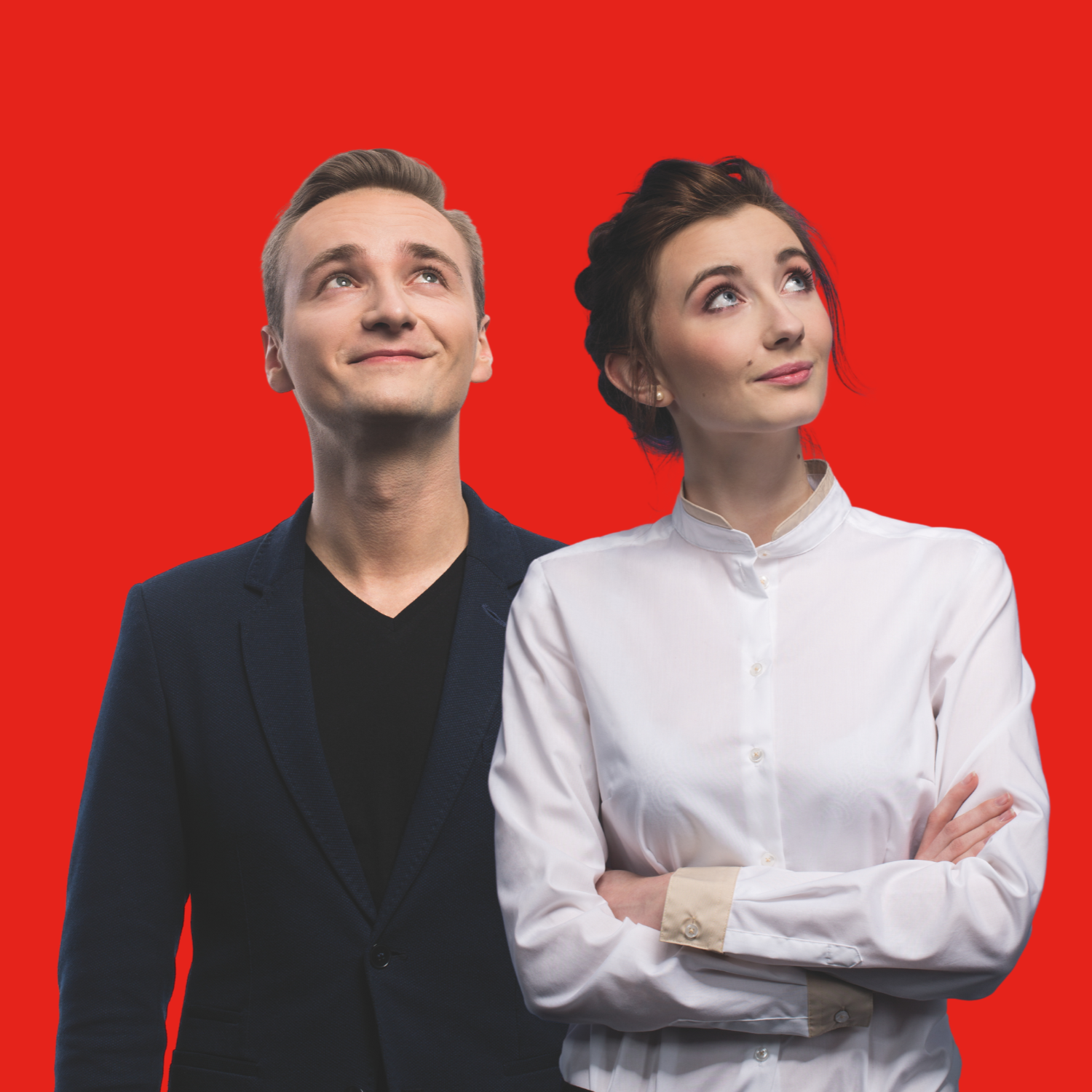 "Młody mężczyzna i kobieta stoją obok siebie na tle intensywnie czerwonego tła, oboje patrzą w górę z lekkimi uśmiechami. Mężczyzna ubrany jest w ciemną marynarkę i czarną koszulkę, a kobieta ma na sobie elegancką, białą koszulę z kołnierzykiem i skrzyżowane ręce."
