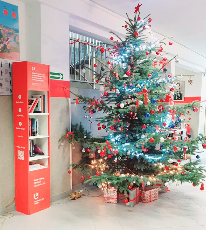 Zdjęcie przedstawia korytarz w szkole. Stoi choinka. Obok niej stoi czerwony regał z książkami. 