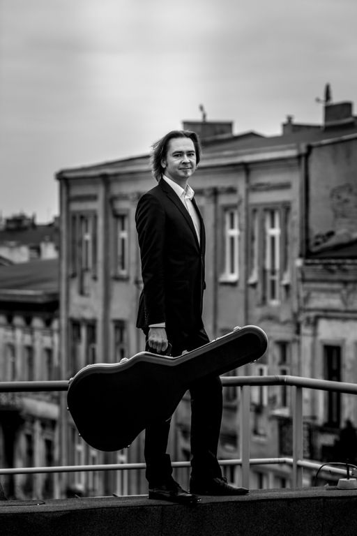 Maciej Staszewski stojący w garniturze z gitarą na balkonie, w tle widoczne są kamienice
